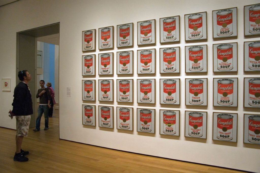 Photo of man observing several side-by-side paintings of various Campbell Soup cans in a museum