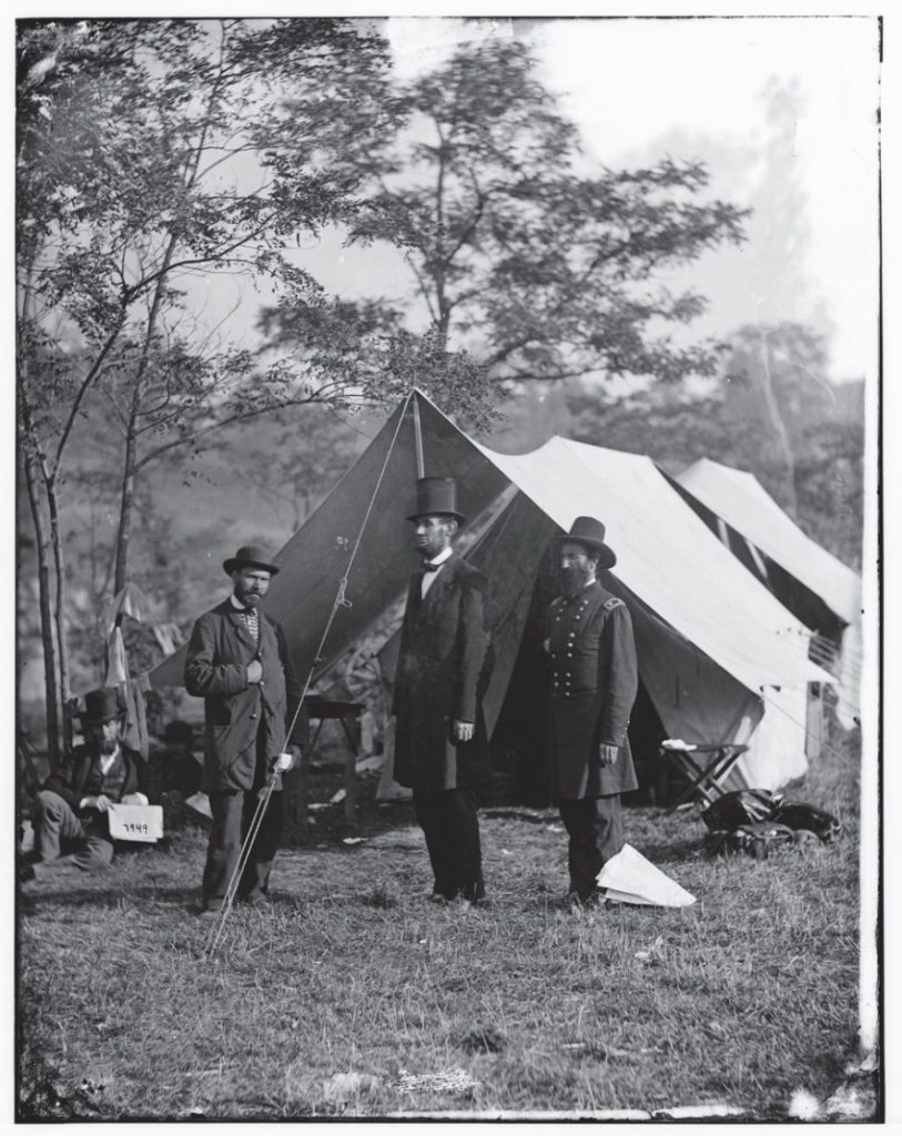 Photo of Abraham Lincoln at center with a man at each side and soldiers' tents behind them.