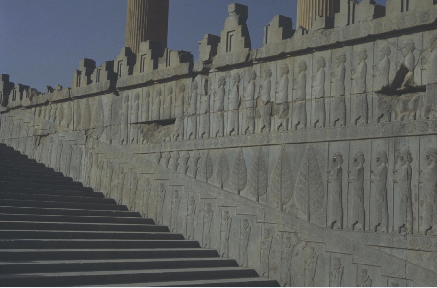 Photo of a stone wall showing figures lined up in orderly, even rows with staircase adjacent.