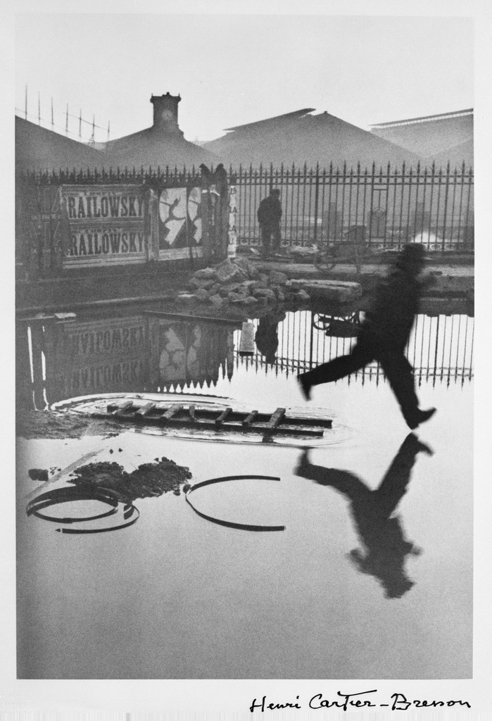 Image of Behind the Gare Saint-Lazare by Henri Cartier-Bresson. Photograph of a scene where a man appears to be leaping, capturing that exact instant.