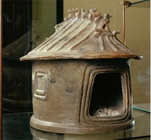 A ceramic urn in the shape of a small house or hut with carved birds on the top.