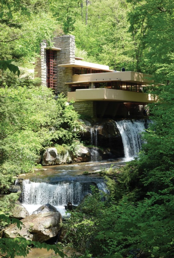 Photo of stone house set into wooded hillside with platforms extending outward and waterfall at bottom.