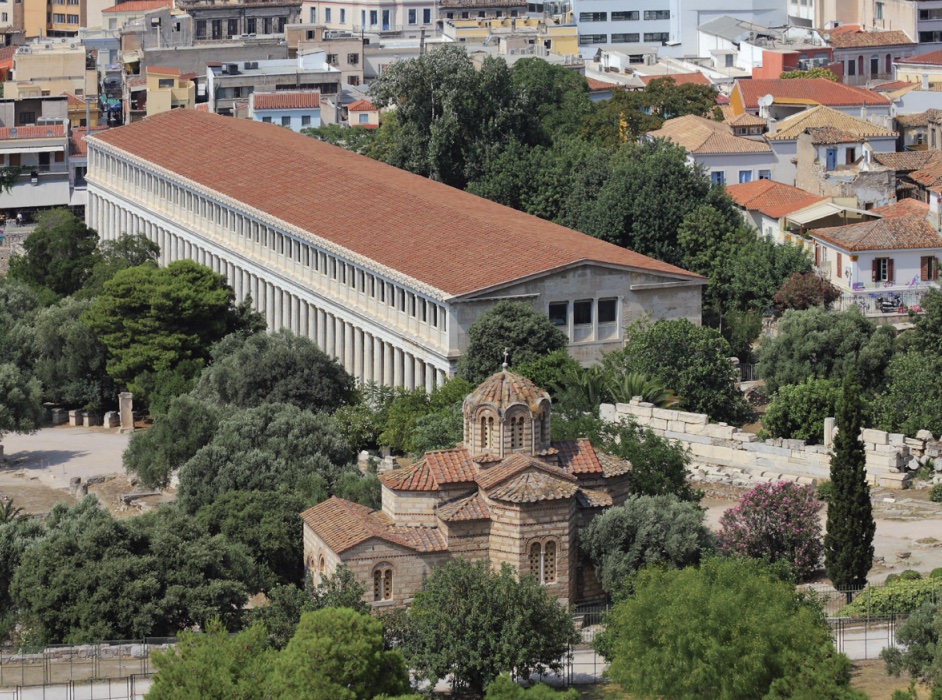Photo of very long building with column along entire length and red roof set among cityscape.