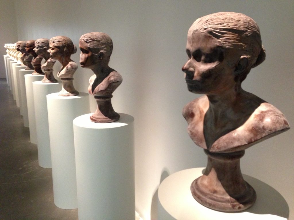 Row of sculpted heads, each on a white pedestal, lined up along a museum wall.
