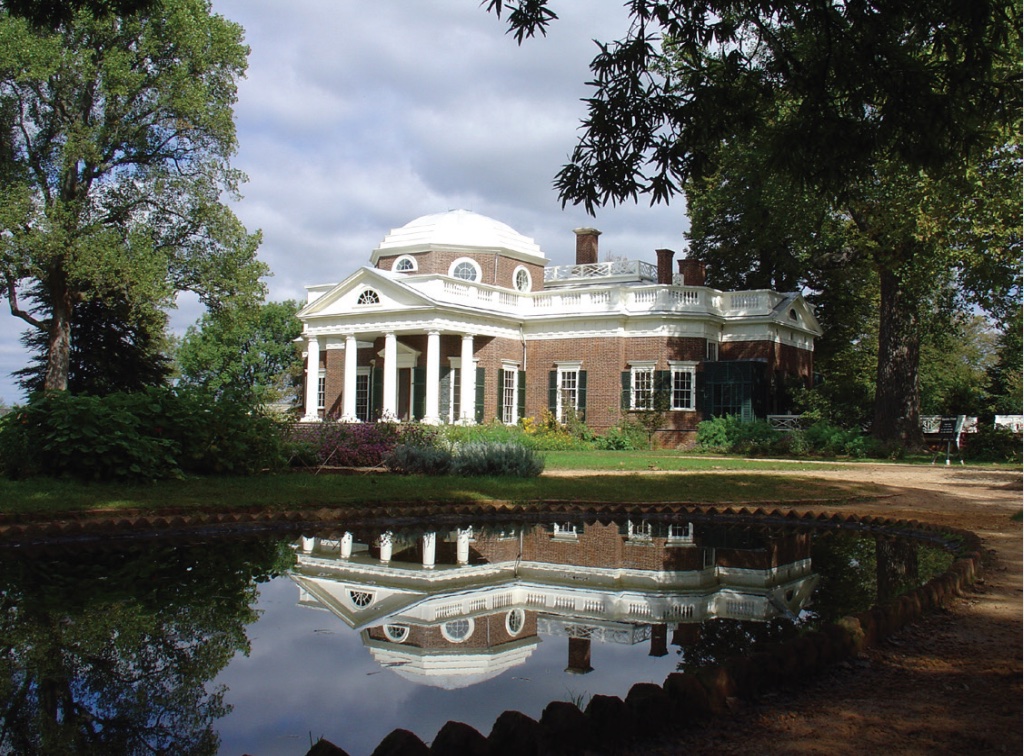 Photo of a brick house with white dome top, white columns, and black shutters around windows set among water and trees.