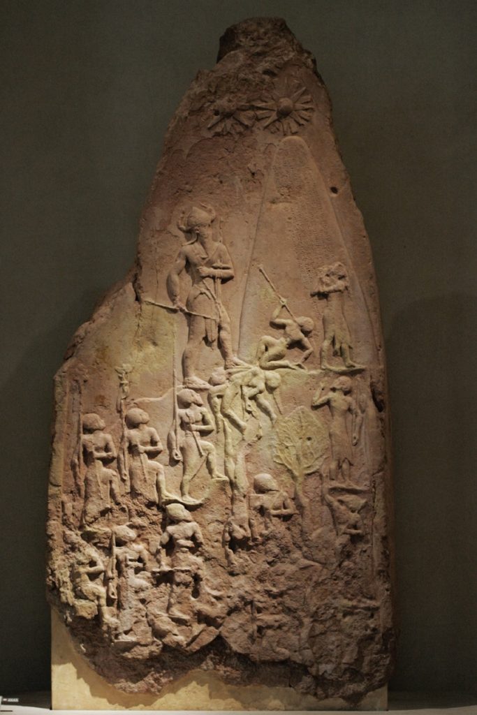Photo of a stele depicting victory of Naram Sin over Lullubi, wearing a horned helmet and appearing much larger than surrounding men.