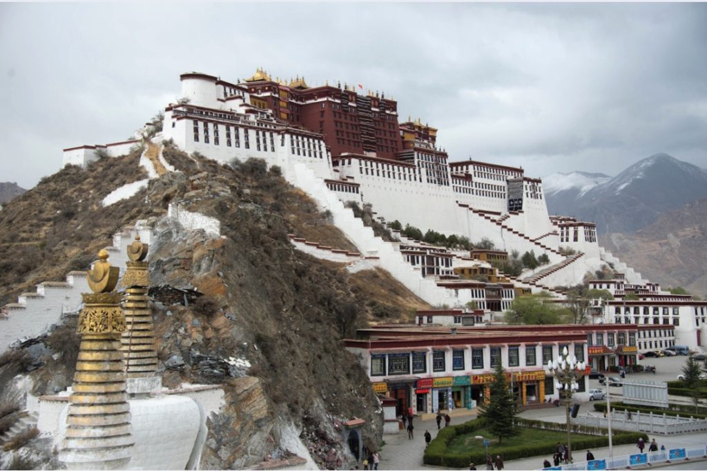 Photo of a white and brown Buddhist palace sitting at top of hill with shops and other village buildings at foot.