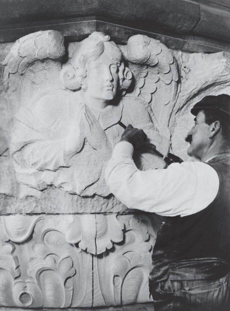 Black and white photo of sculptor in vest and cap sculpting an angel in stone.