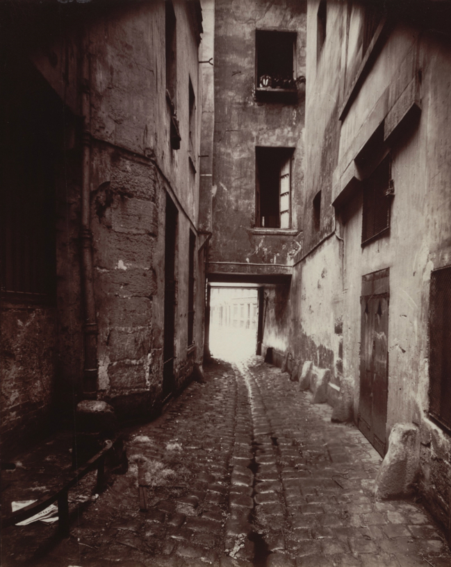 Courtyard, 22 rue Quincampoix by Eugène Atget. Photograph of a courtyard where windows to homes can be seen, the way out from the courtyard can be seen.