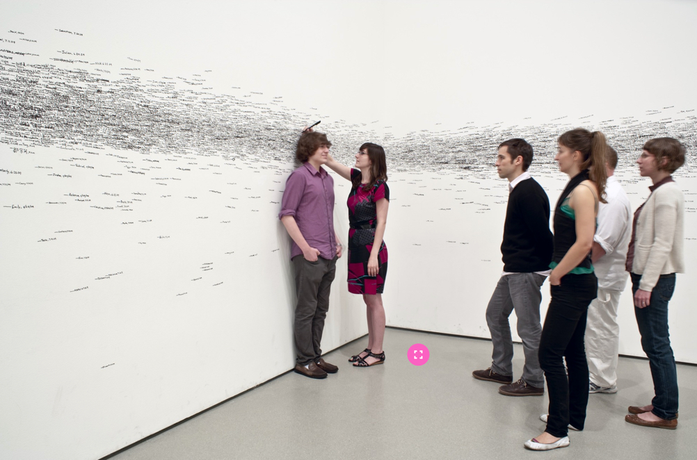 Image of Measuring the Universe, on display by Roman Ondák. Large white wall room with black marks going all around the room. A crowd of people is visible looking at one of the walls.