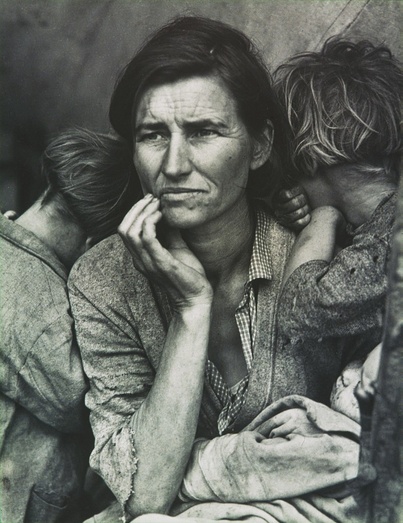 Photograph of a mother with a dirty face holding her two children inside of a tent.