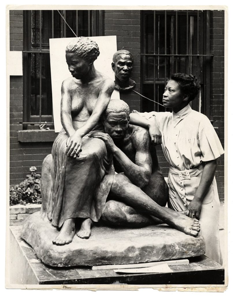 Artist standing beside her sculpture of a woman seated in a chair topless in a long skirt, and a man curled, nude beside her sitting on the ground with his hand in her lap.