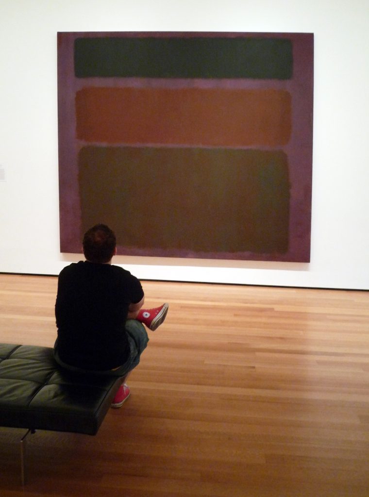 Image of No.16 (Red, Brown and Black) by Mark Rothko on display. The painting depicts three rectangular shapes starting with a slimmer rectangle at the top a bigger rectangle in the middle and an even wider rectangle at the bottom. A man sits in front of the painting on display.