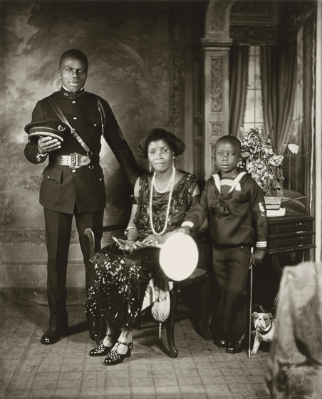 A photo of a family in an elegant sitting room, father in a police or military uniform standing, mother seated in a dress and long pearls, and a son in a sailor suit standing beside a toy dog or puppy.