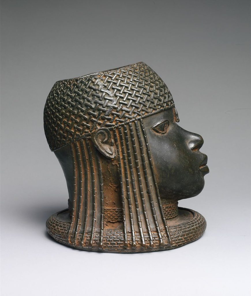 Image of Head of an Oba. Brass head figure with strong realistic features of the face. Wearing a woven cap of coral beads with strands coming down on the sides, also wearing a rolled collar.