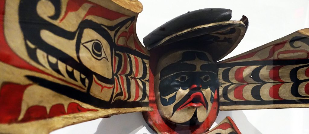 An inuit mask depicing a mans masked face in the center and two large wings with petroglyphs painted on them.