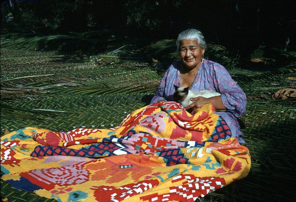 Photo of a woman sitting on the ground, holding a cat, and sewing a patchwork quilt.