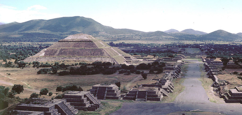 A large stone temple in the shape of a pyramid. A street lined with the ruins of smaller pyramids is in the foreground.