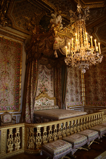 Image of the Queen's bed chamber at Versailles. A huge chandelier hangs over the bed which has a gold railing in front.