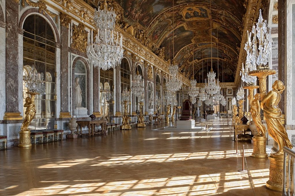 Image of the Hall of Mirrors in the Palace of Versailles. Chandeliers hang throughout the hall with various gold Sculptures at the sides.