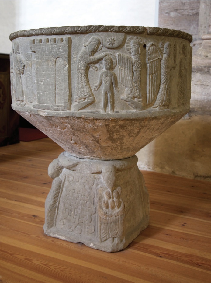 Photo of stone baptismal font with figures carved into side depicting a baptism.