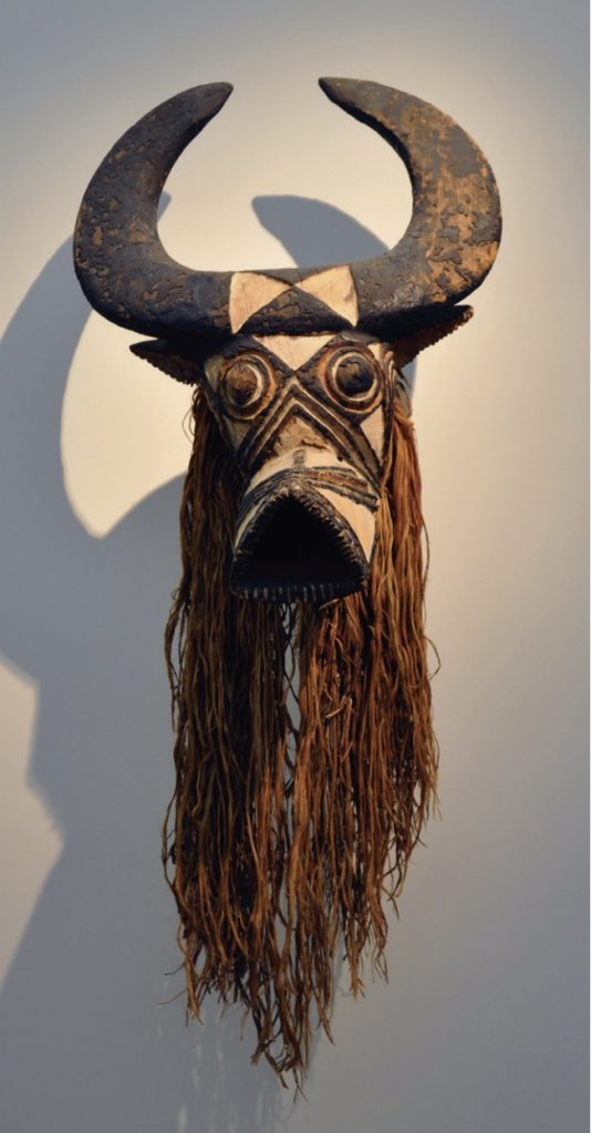 Photo of mask with animal head, large horns, long hair-like material along sides.