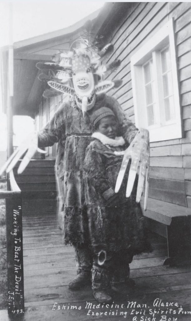 Black and white photo of Eskimo medicine man in traditional dress with mask posing with small child.