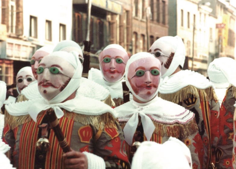 Photo of people in masks with painted faces and colorful costumes parading down street in city.