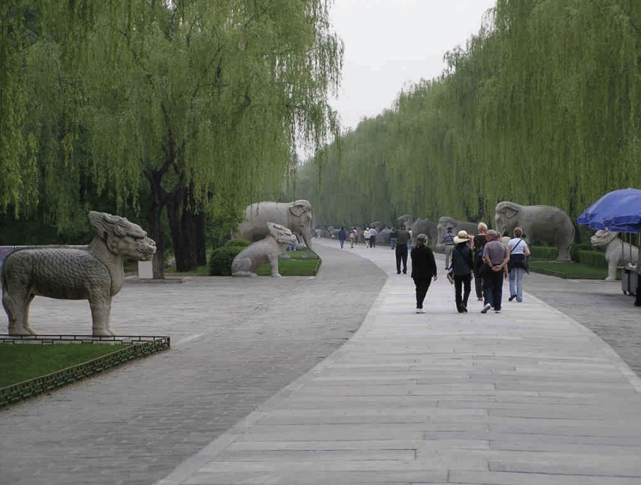 Photo of a concrete path lined with statues and trees with people observing along the length.