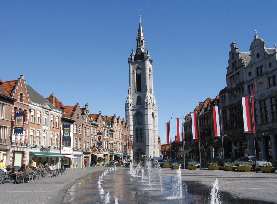 Photo of a grey steeple with city street, shops, and fountains in foreground.
